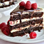 A slice of chocolate cake layered with white frosting and topped with fresh raspberries and chocolate ganache sits on a white plate.